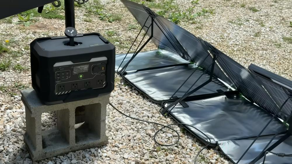 A portable black power station sits on a cinderblock outdoors, connected to a folded black solar panel setup on gravel ground.