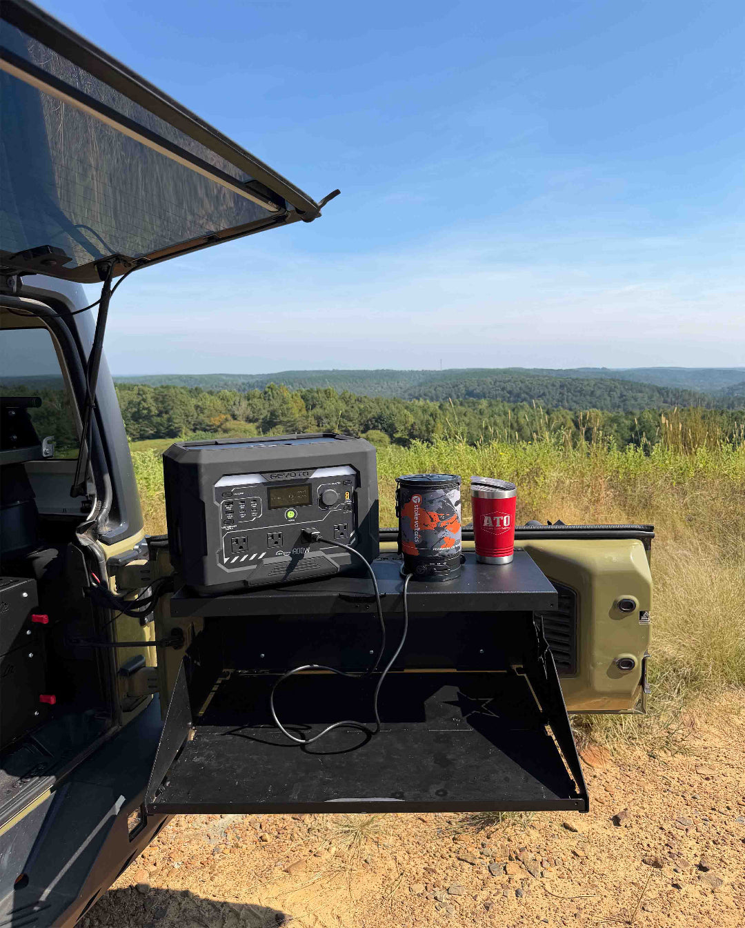 An outdoor camping setup with a portable power station and two insulated tumblers placed on a metal shelf extended from the side of a vehicle, with a scenic forest landscape in the background.