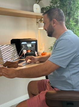 A man wearing a light blue t-shirt and pink shorts sits in a home office environment, working at a wooden desk with a laptop, 3D printer, and phone. The background features light wood floating shelves, a green houseplant, and a window.