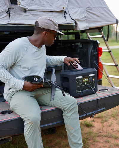 A person seated on a pickup truck tailgate beside a GEYOTO power station and connected devices at a campsite.
